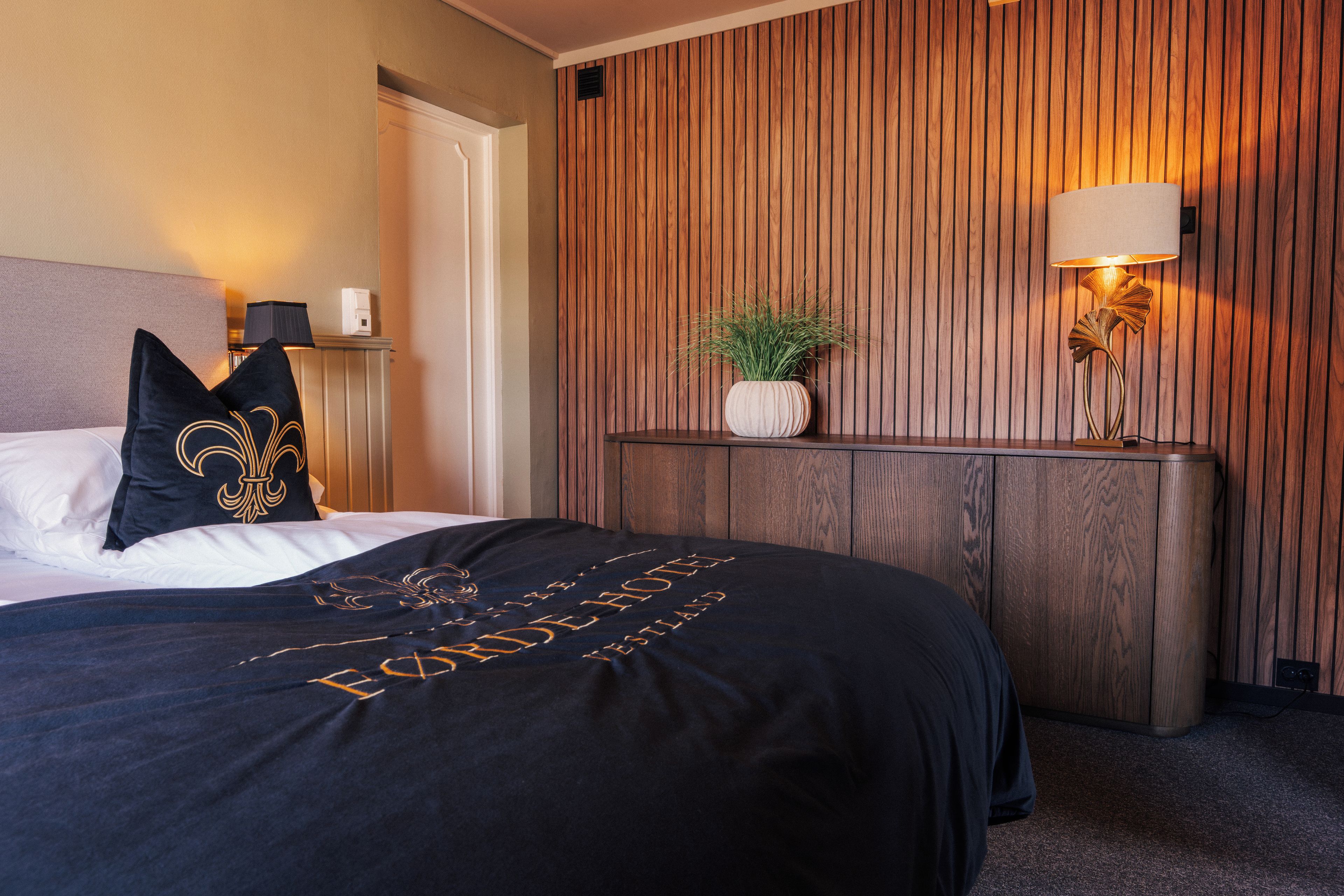 A hotel room with a bed, a nightstand, a lamp, and a wall with vertical wooden panels.