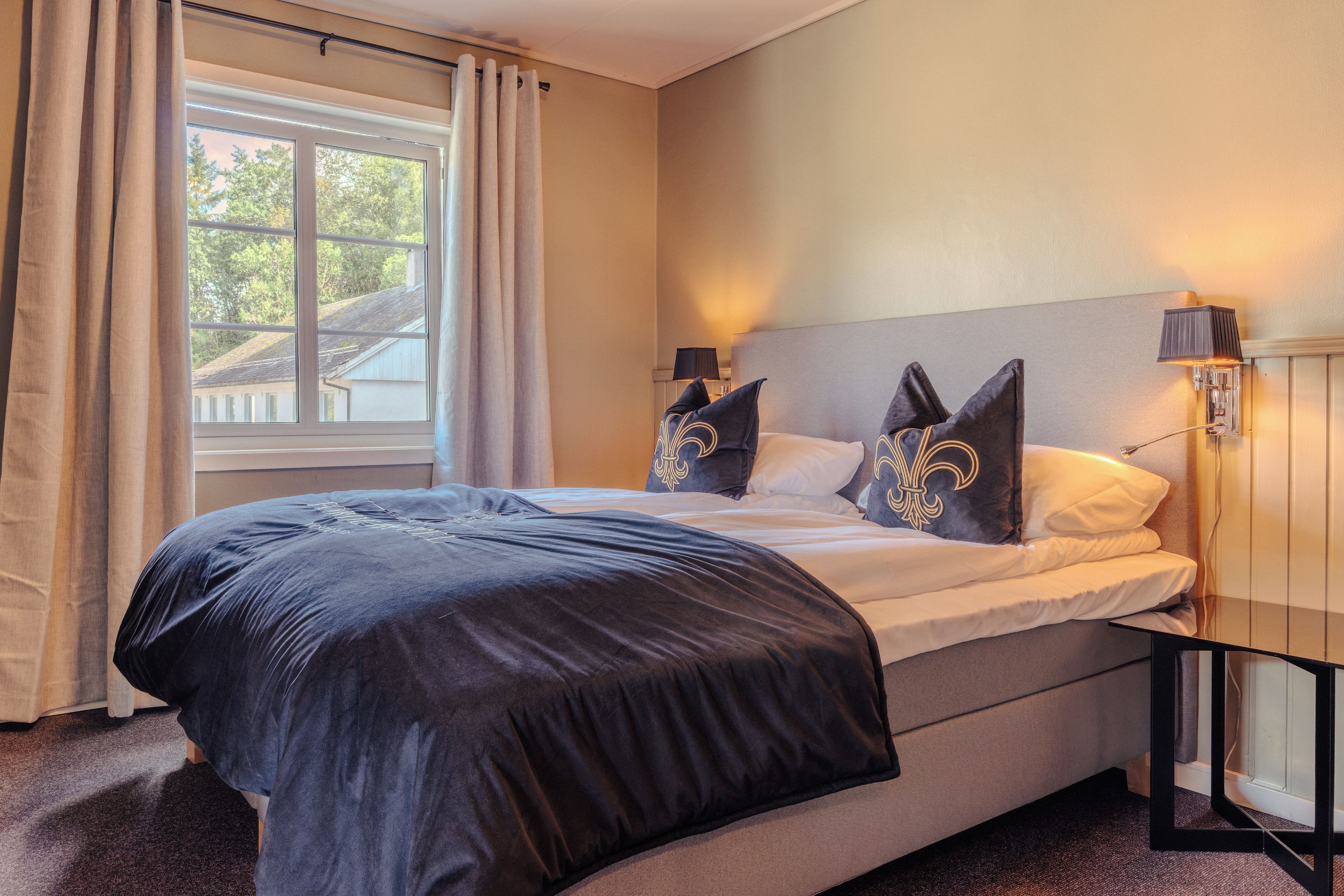 A hotel room with a bed, a window with curtains, a nightstand with a lamp, and a wall-mounted light fixture.