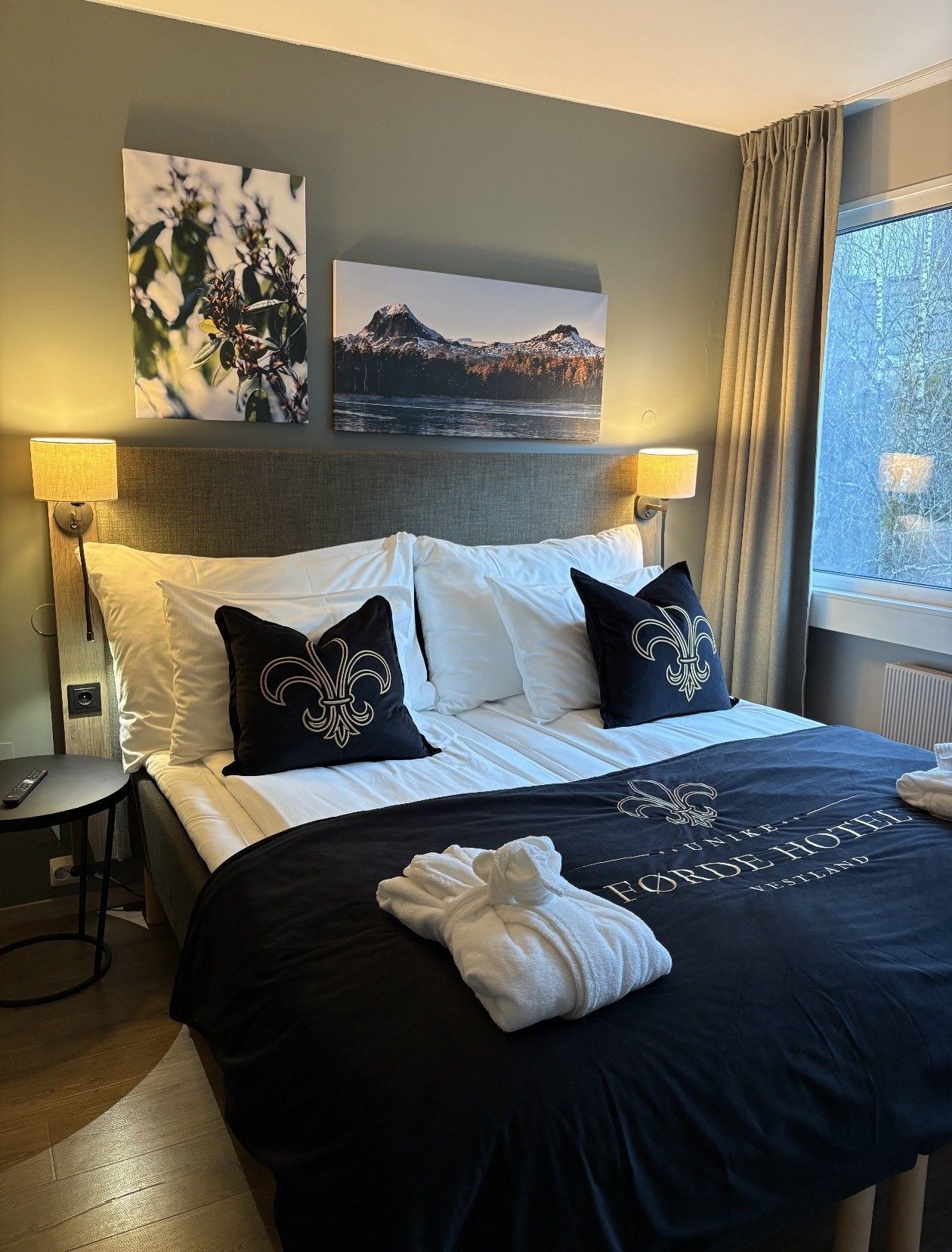A hotel room with a bed, two bedside lamps, a nightstand, a remote control, and a window with curtains.