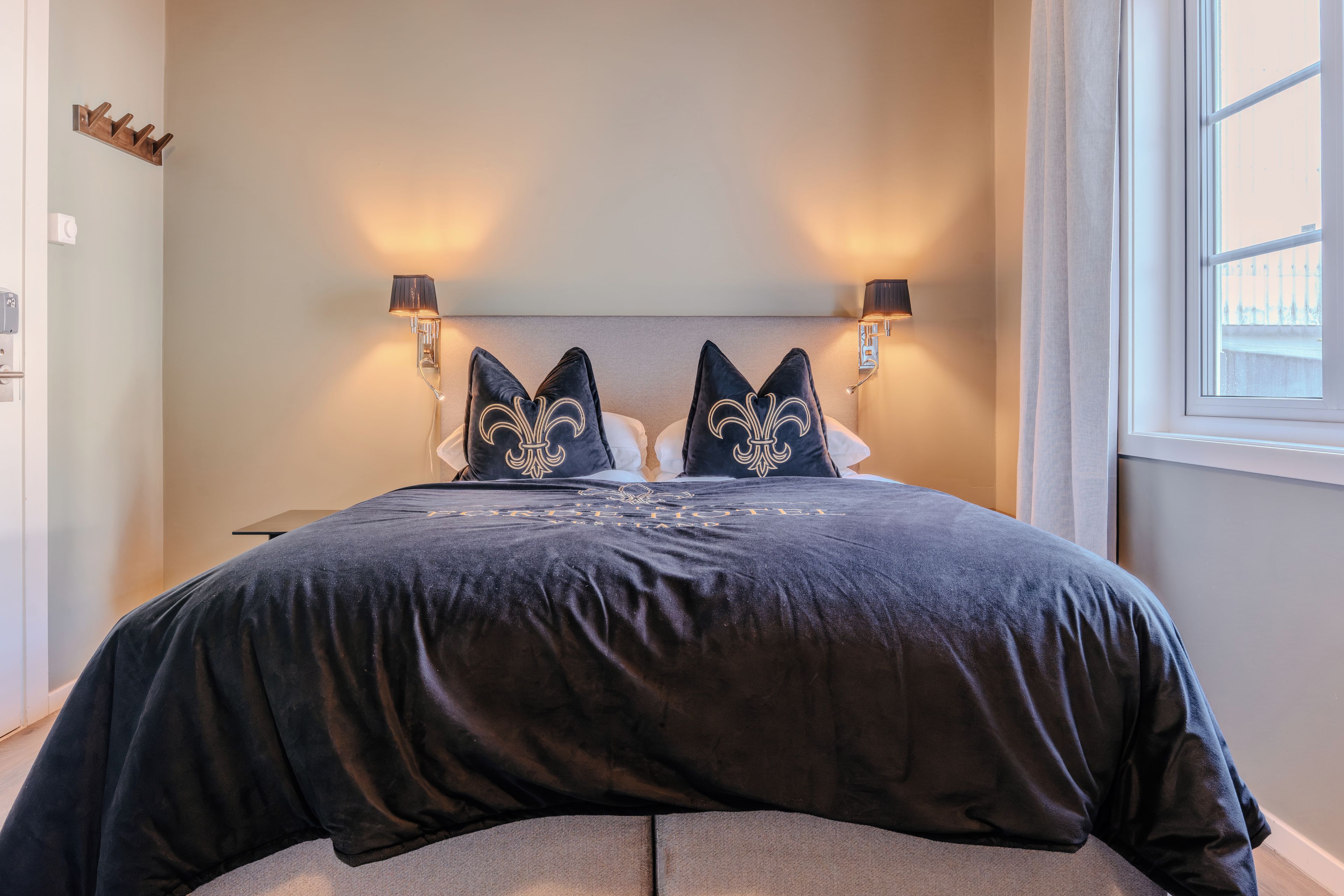 Uma cama com colcha escura e duas almofadas decorativas, uma mesa de cabeceira com abajur, e uma janela com cortinas.