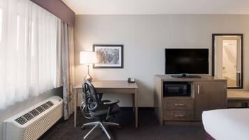 A hotel room with a bed, a desk with a chair, a television, a microwave, a heater, a window with curtains, and a framed picture on the wall.