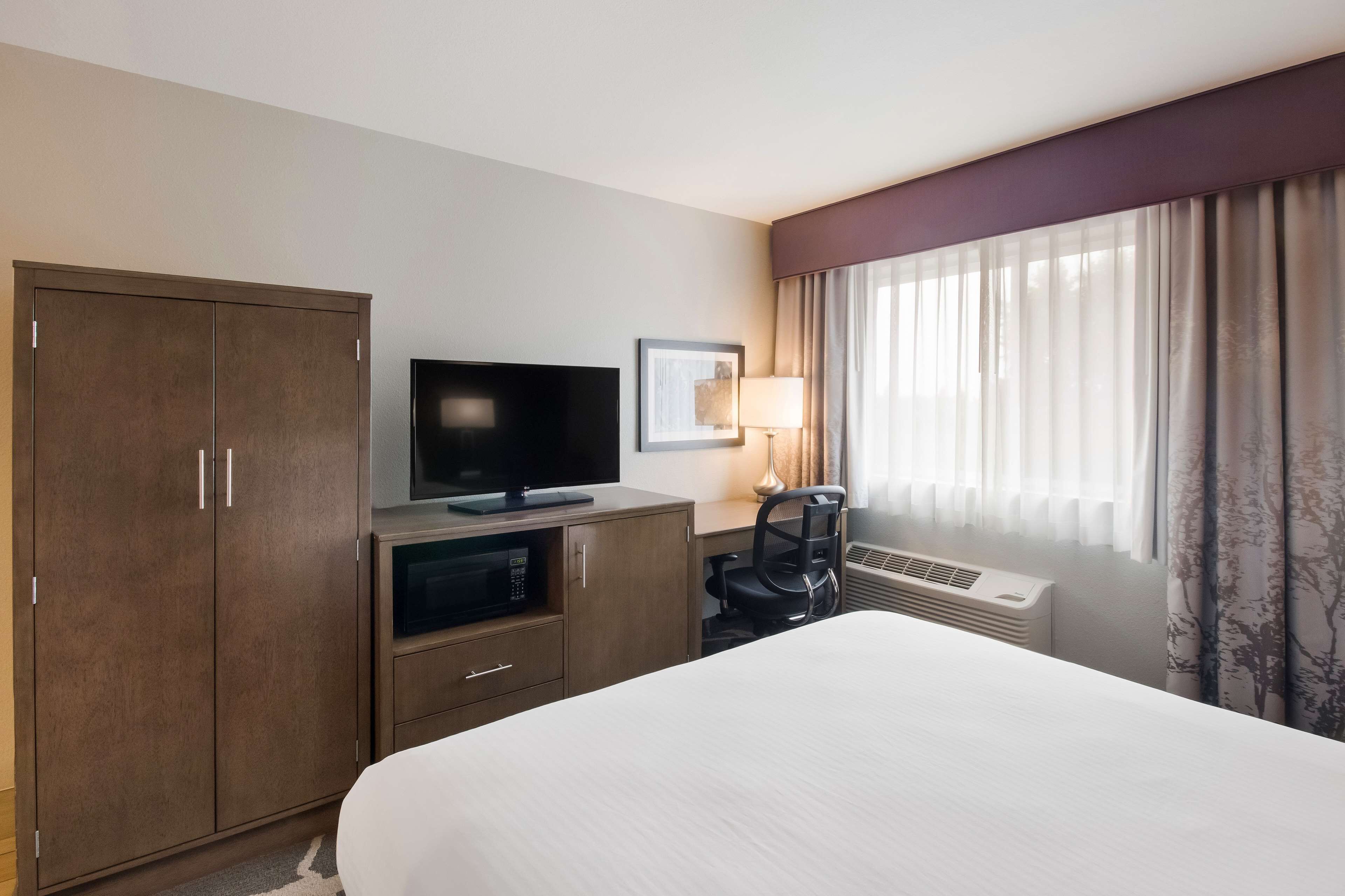 A hotel room with a bed, a TV, a desk with a lamp, a chair, and a wardrobe.