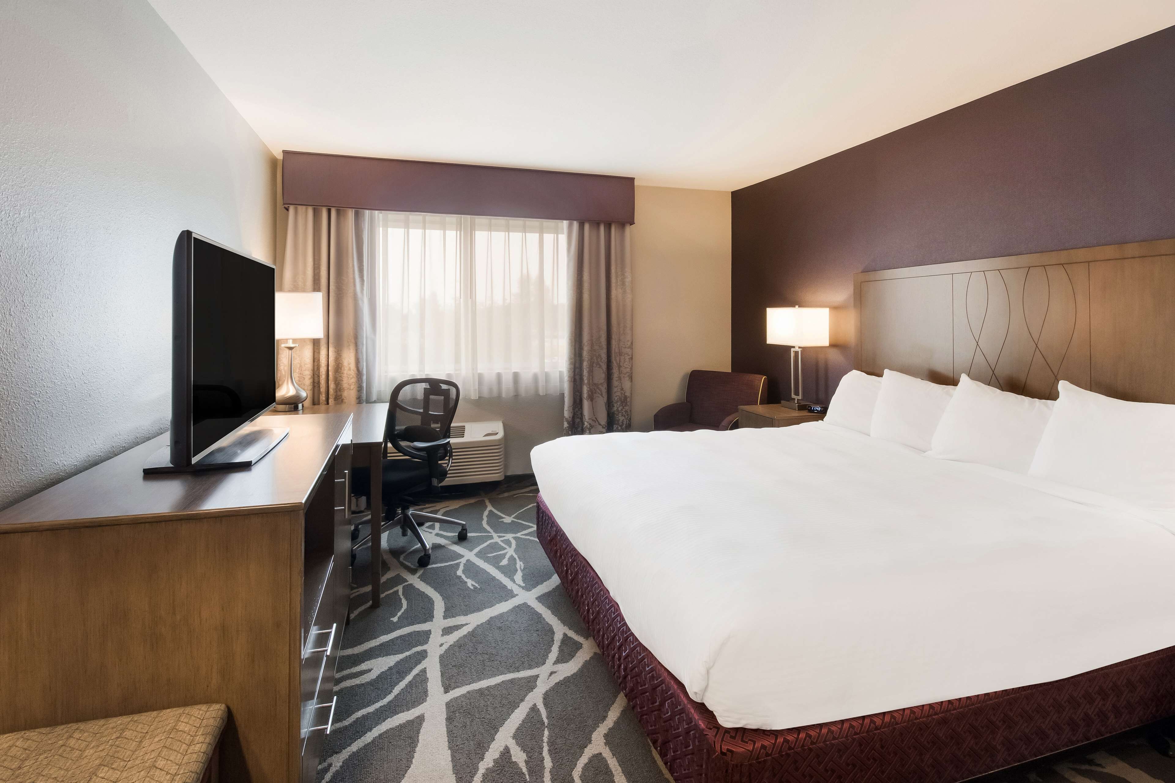 A hotel room with a large bed, a desk with a flat-screen TV, a chair, and a lamp.