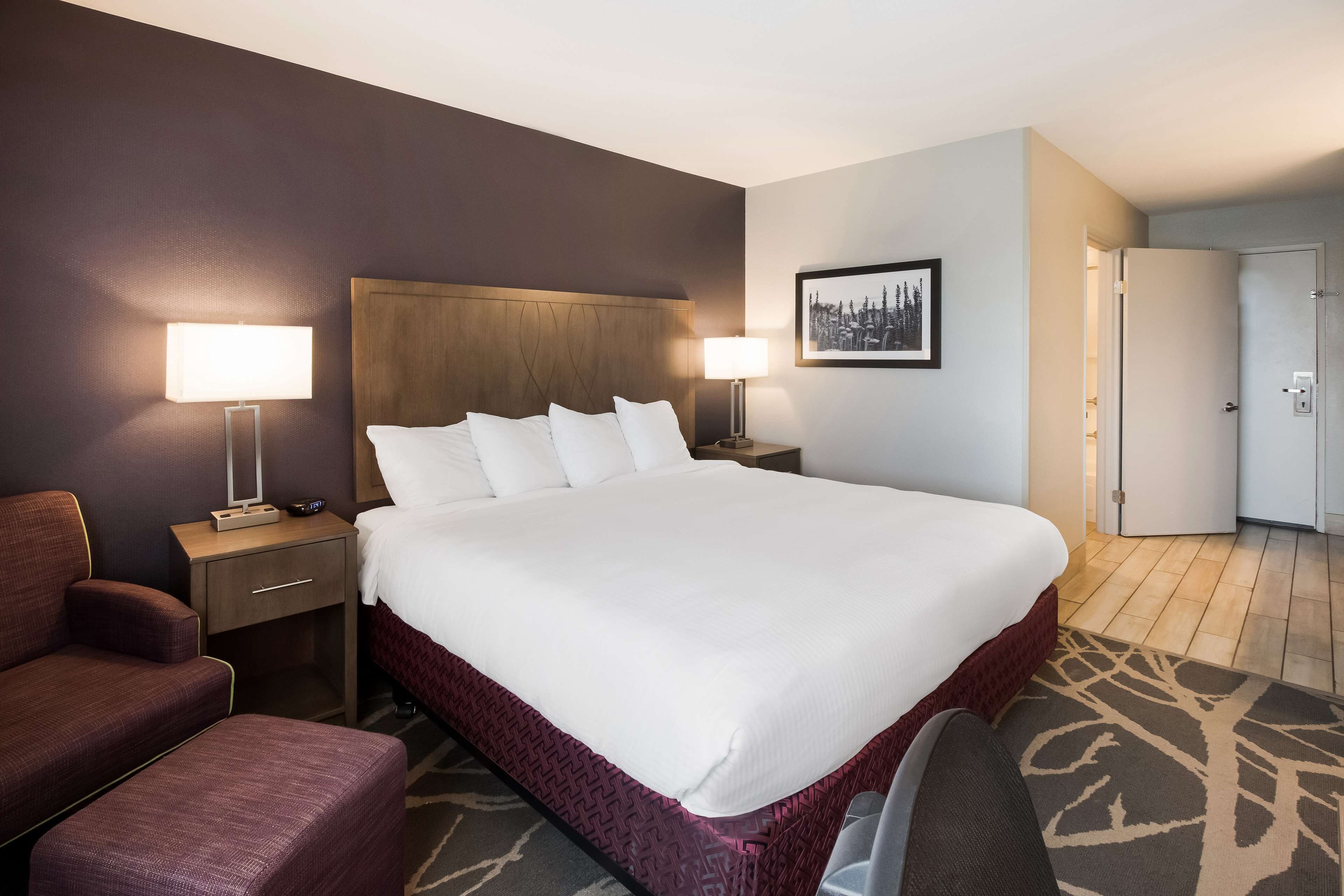 A hotel room with a large bed, two bedside tables with lamps, a chair, and a door leading to another room.