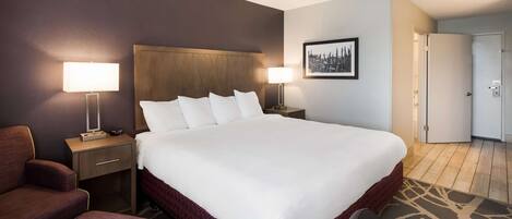 Habitación de hotel con una cama grande, dos mesitas de noche con lámparas, una silla y una puerta que da a otra habitación.