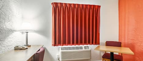 A compact room with a wooden desk, a chair, a table, a lamp, and a window with red curtains.