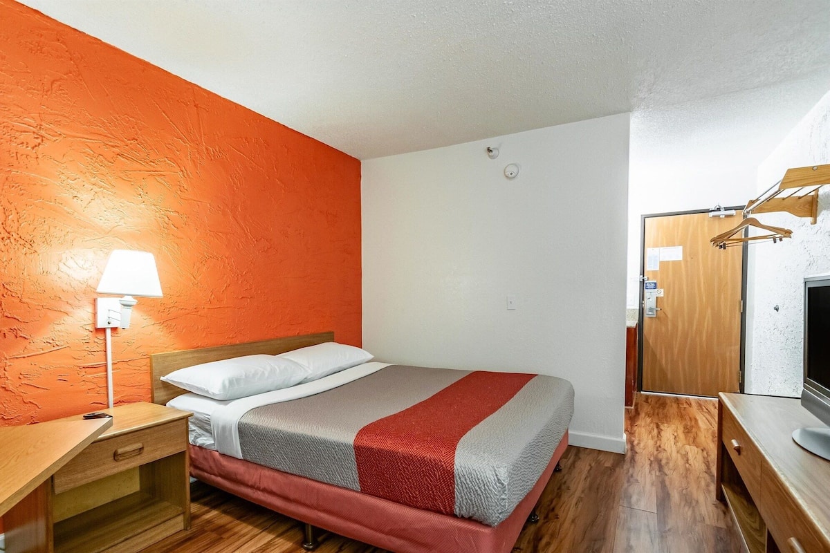 A bedroom with a bed, a nightstand, a lamp, a wooden desk, a TV, and a wardrobe.
