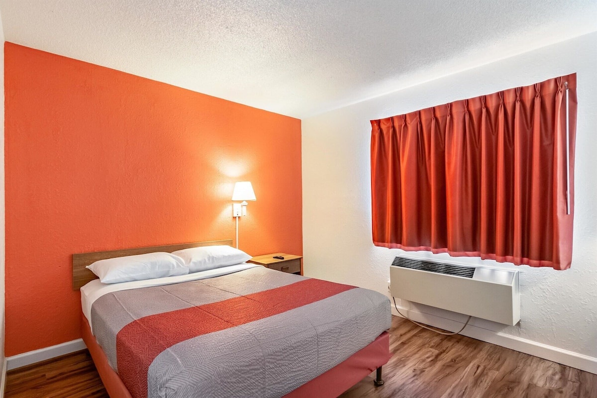 A hotel room with a bed, a nightstand, a lamp, a window with curtains, and an air conditioning unit.