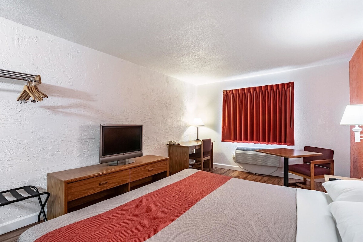 A hotel room with a bed, a desk, a television, a lamp, and a window with red curtains.