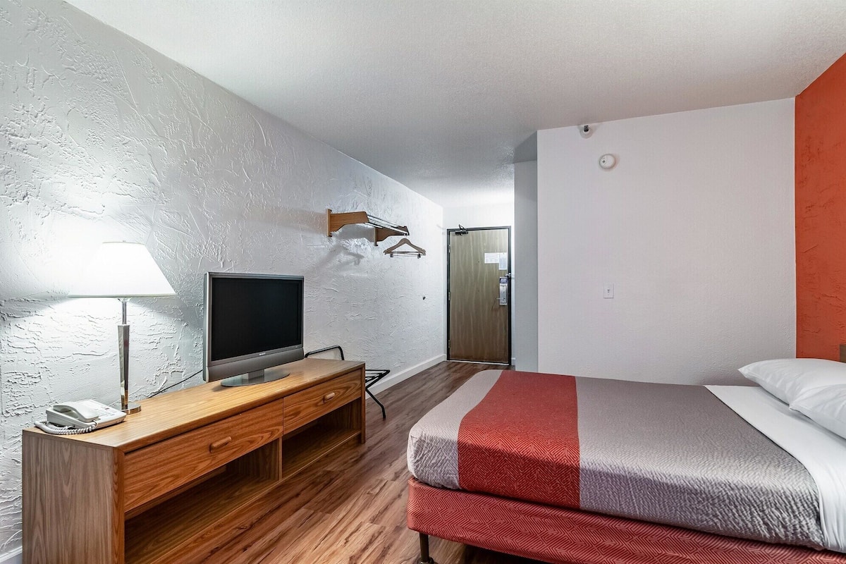 A hotel room with a wooden desk, a flat-screen TV, a bed with red and grey bedding, and a white textured wall.