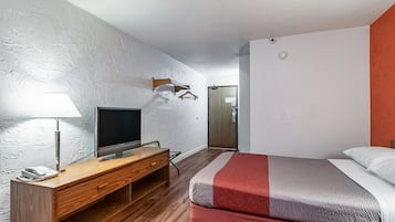 A hotel room with a wooden desk, a flat-screen TV, a bed with red and grey bedding, and a white textured wall.