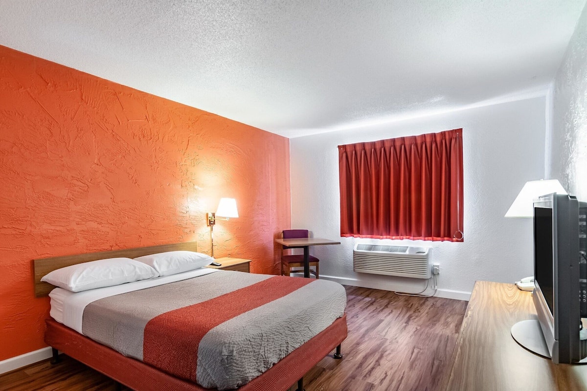 A hotel room with a bed, a nightstand, a lamp, a window with red curtains, an air conditioning unit, and a TV on a wooden table.