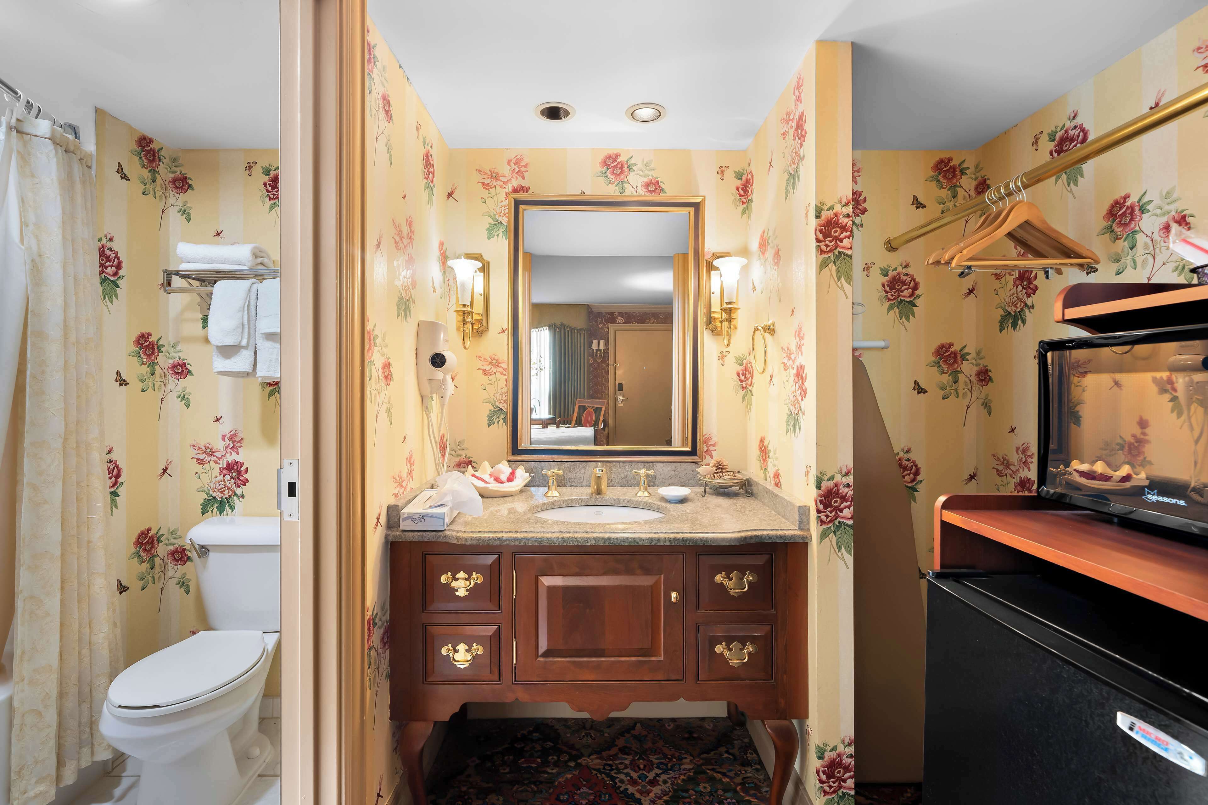 A bathroom with floral wallpaper, a white toilet, a sink with a mirror, and a black refrigerator.