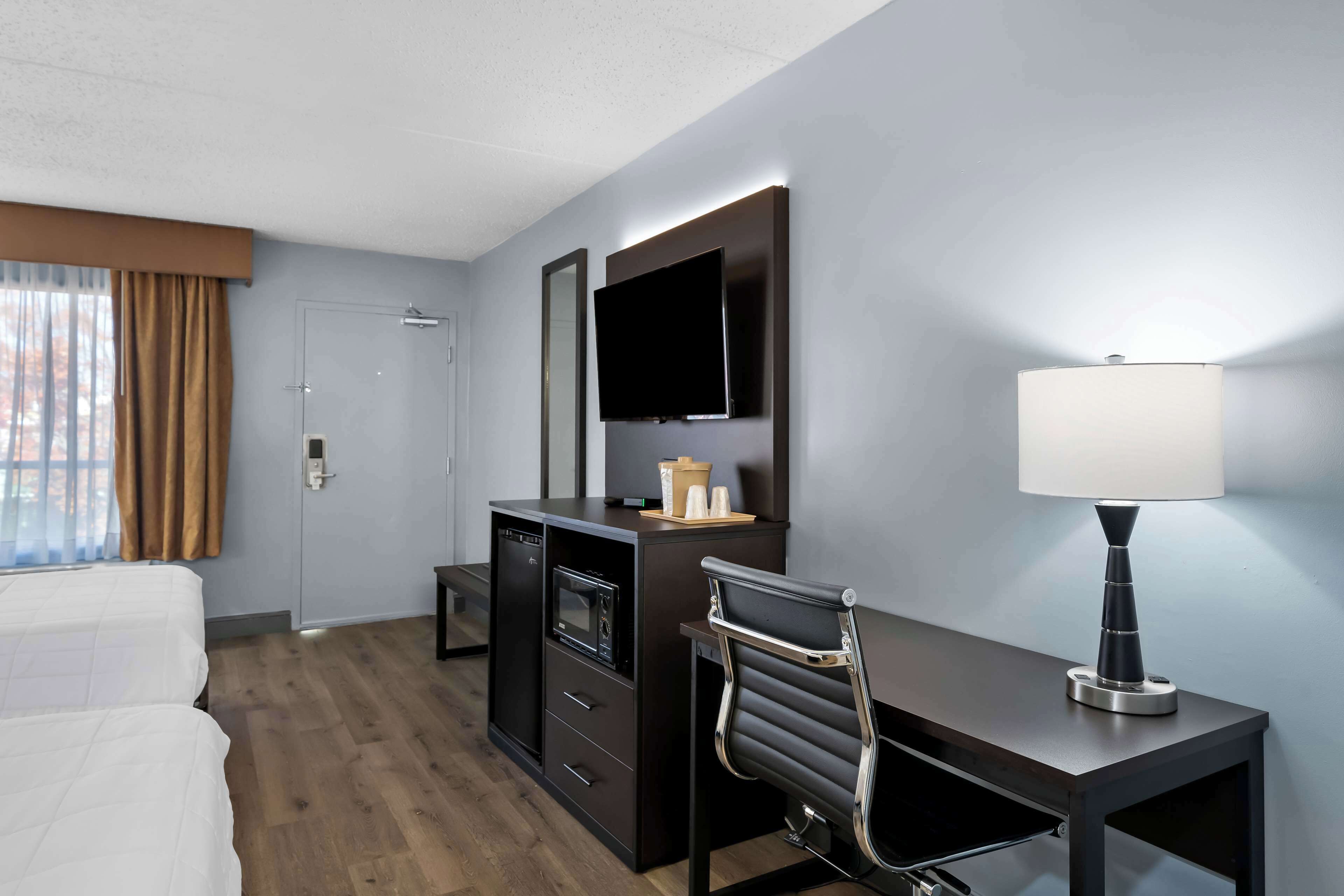 A hotel room with a bed, a desk, a chair, a TV, a lamp, and a minibar.