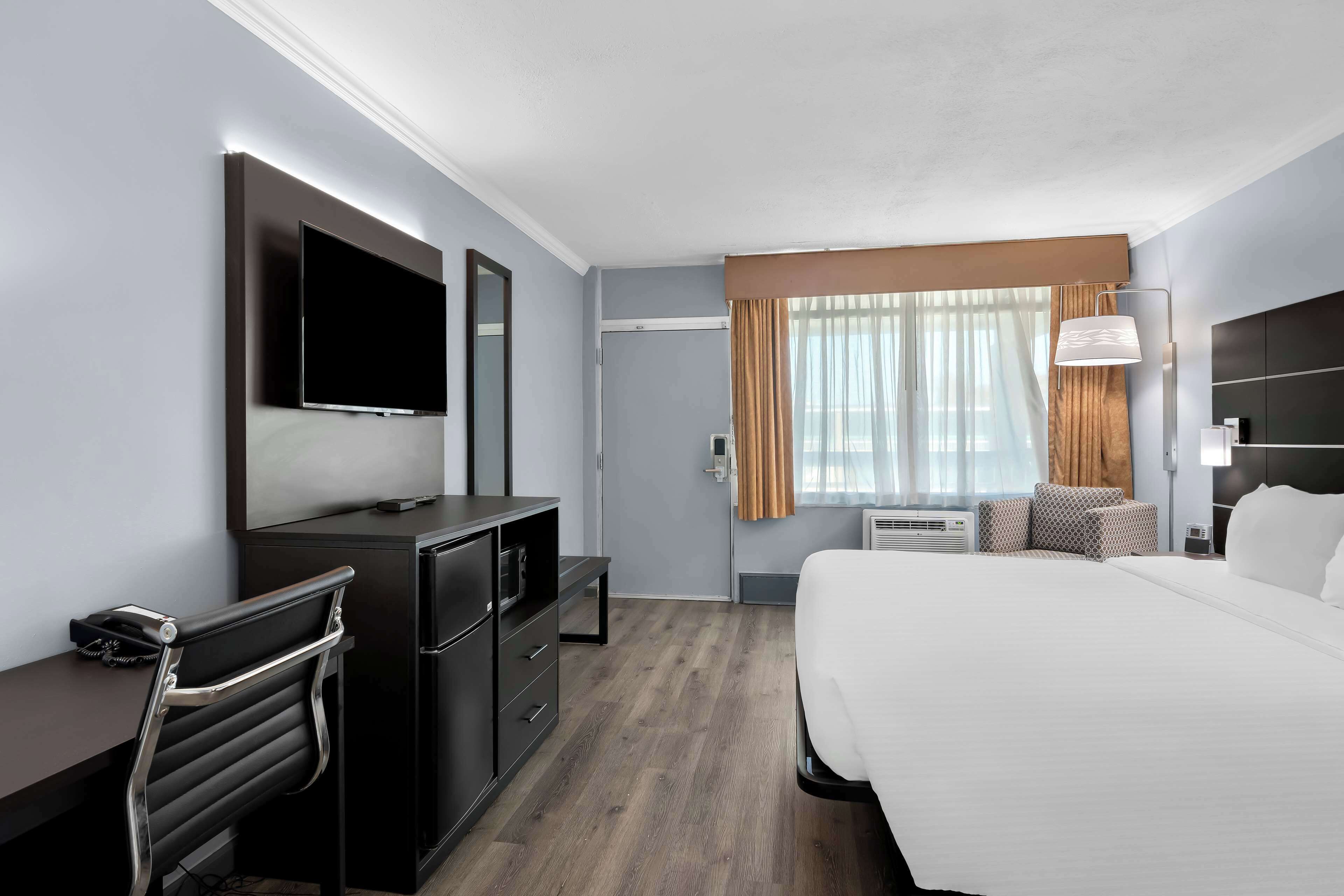A modern hotel room with a large bed, a desk with a flat-screen TV, a dresser, and a seating area with a chair and ottoman.