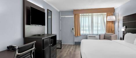 A modern hotel room with a large bed, a desk with a flat-screen TV, a dresser, and a seating area with a chair and ottoman.