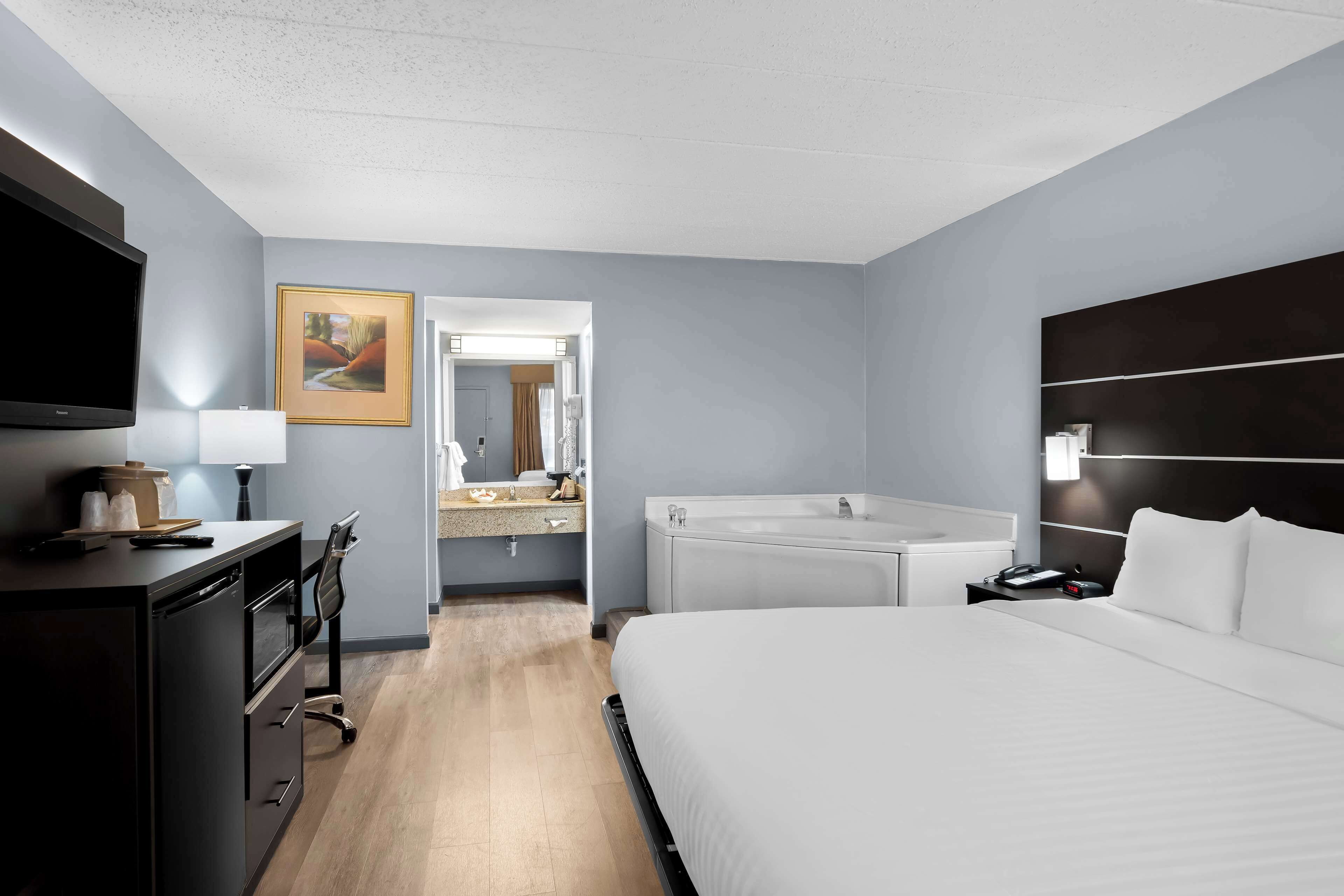 Una habitación de hotel moderna con una cama grande, un escritorio con una computadora, un televisor de pantalla plana, un baño con bañera y un cuadro en la pared.