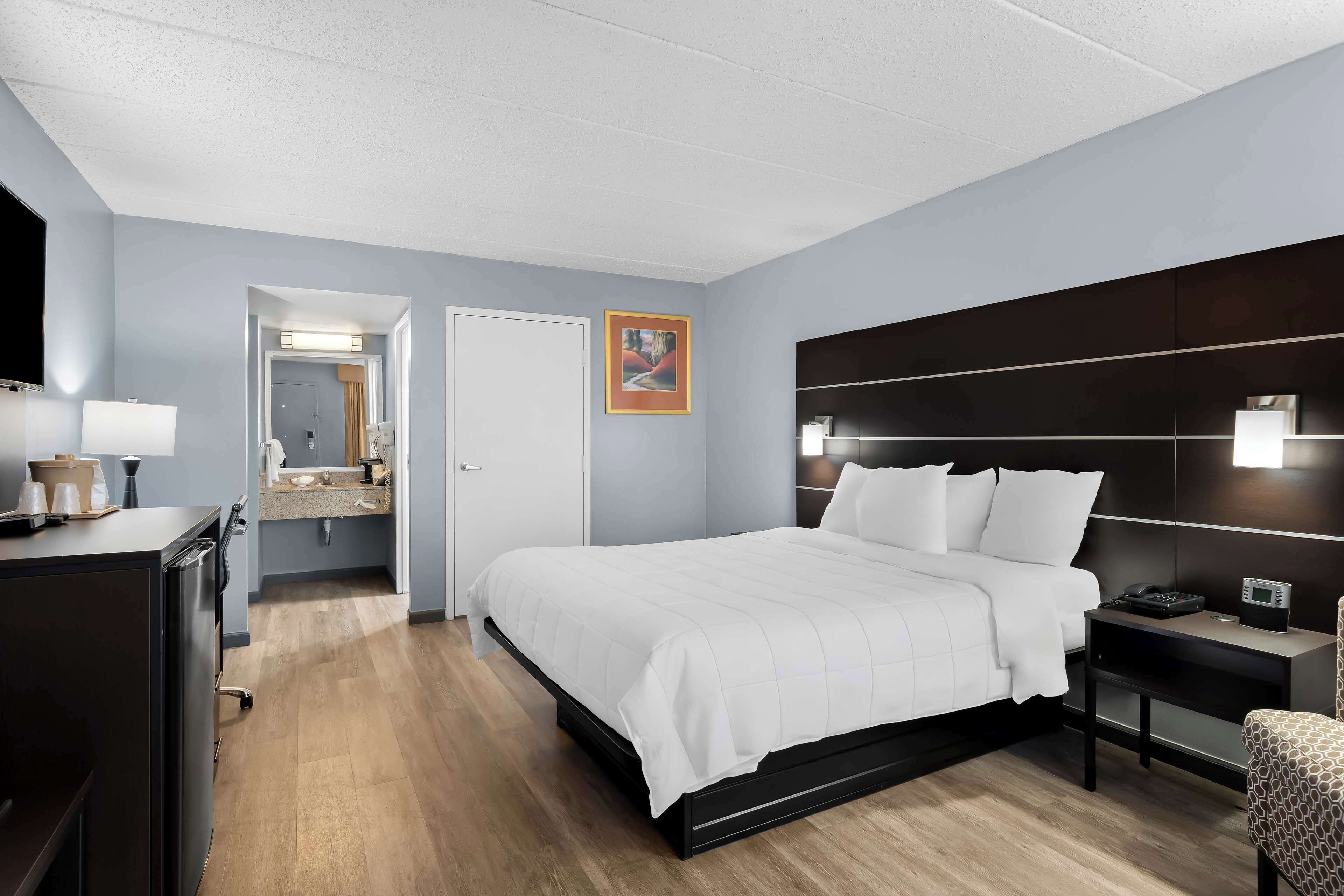 A modern hotel room with a large bed, a bedside table with a lamp, a TV, and a bathroom visible through an open door.