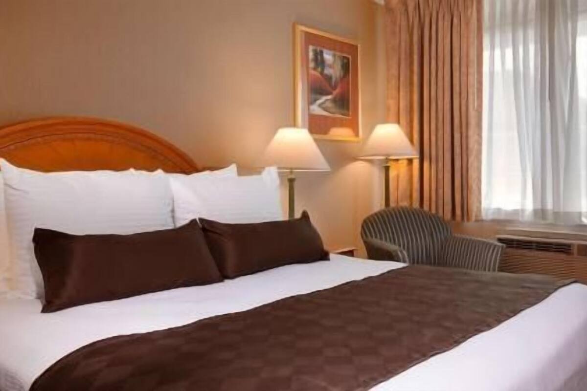 Une chambre d’hôtel avec un lit, deux lampes de chevet, une chaise, un tableau au mur et une fenêtre avec des rideaux.