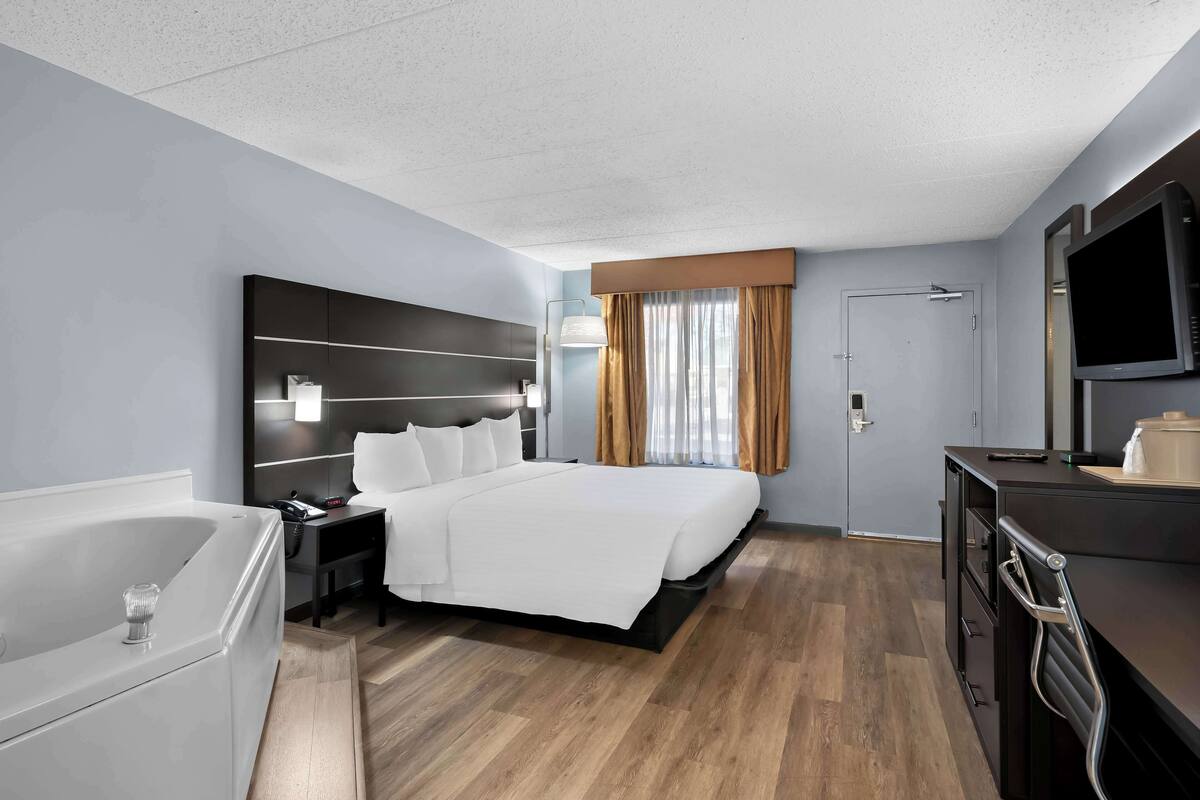 Une chambre d’hôtel avec un grand lit, un bureau avec une télévision, une lampe de bureau, une table de chevet et une salle de bain avec une baignoire, ainsi qu’une porte munie d’un rideau.