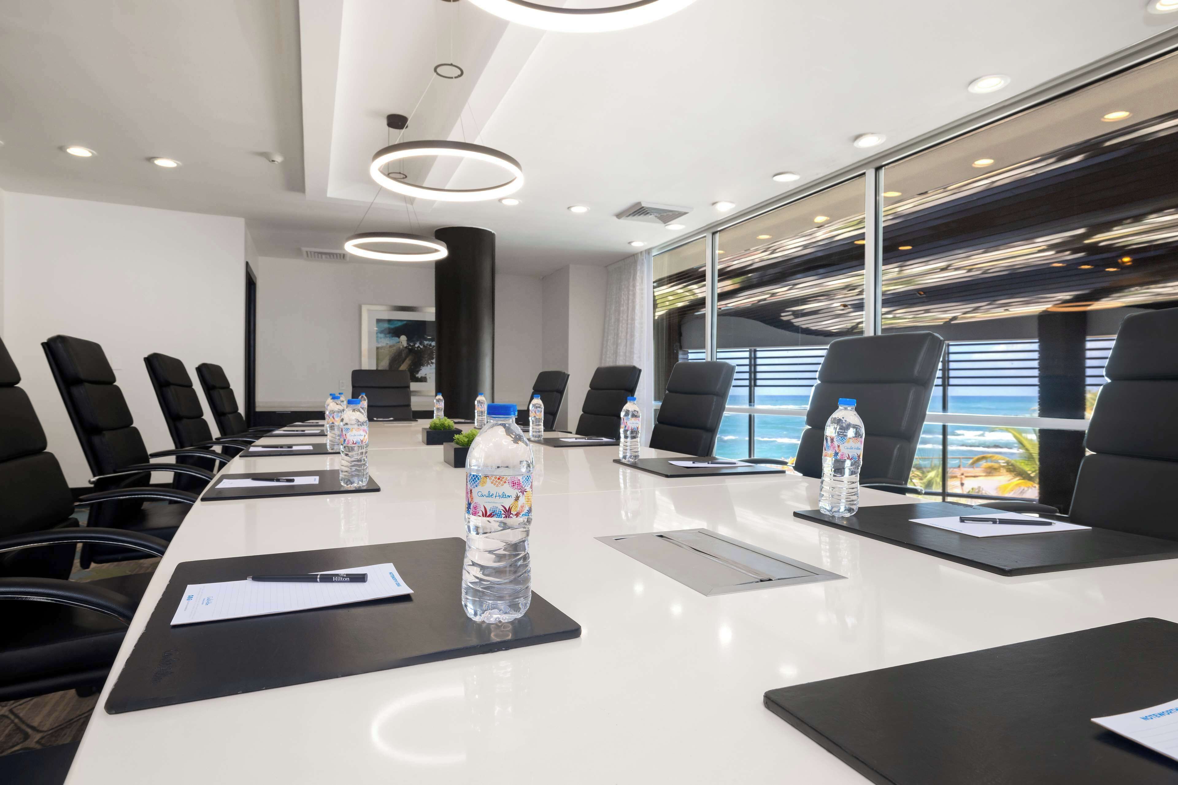 A modern conference room with a long table, black chairs, water bottles, and a view of the ocean through large windows.