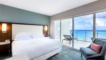 A hotel room with a large bed, a chair, a desk, and a balcony with a view of the ocean.
