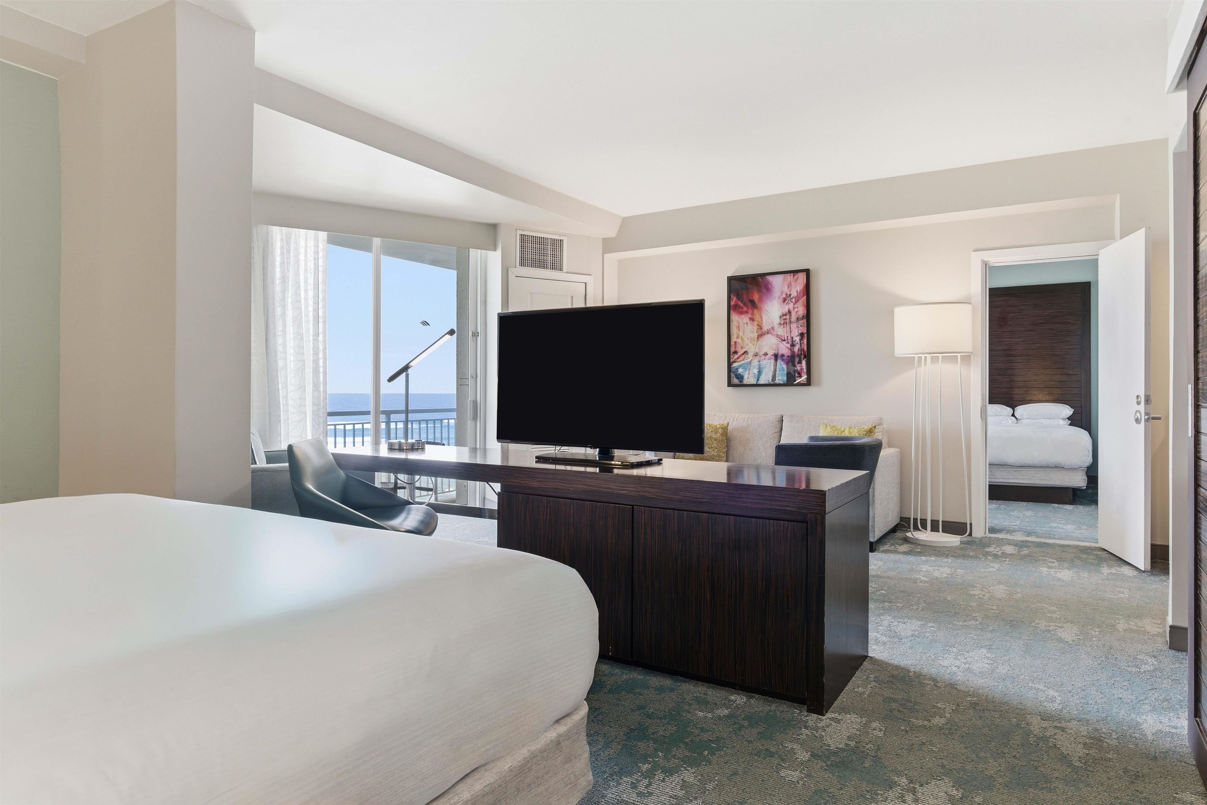 A hotel room with a large bed, a desk with a flat-screen TV, a seating area with a chair, a painting on the wall, and a view of the sea through the window.