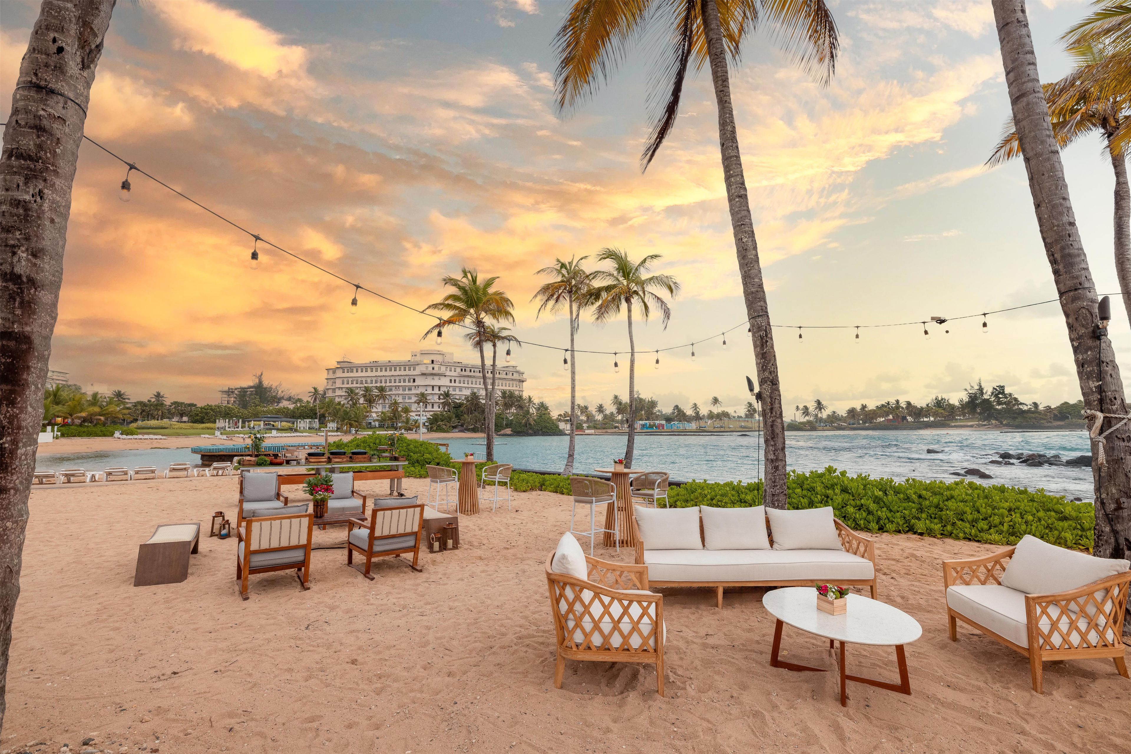 Eine Strandlounge mit Sitzgelegenheiten im Freien, Palmen und Blick auf das Meer sowie ein Hotel im Hintergrund.