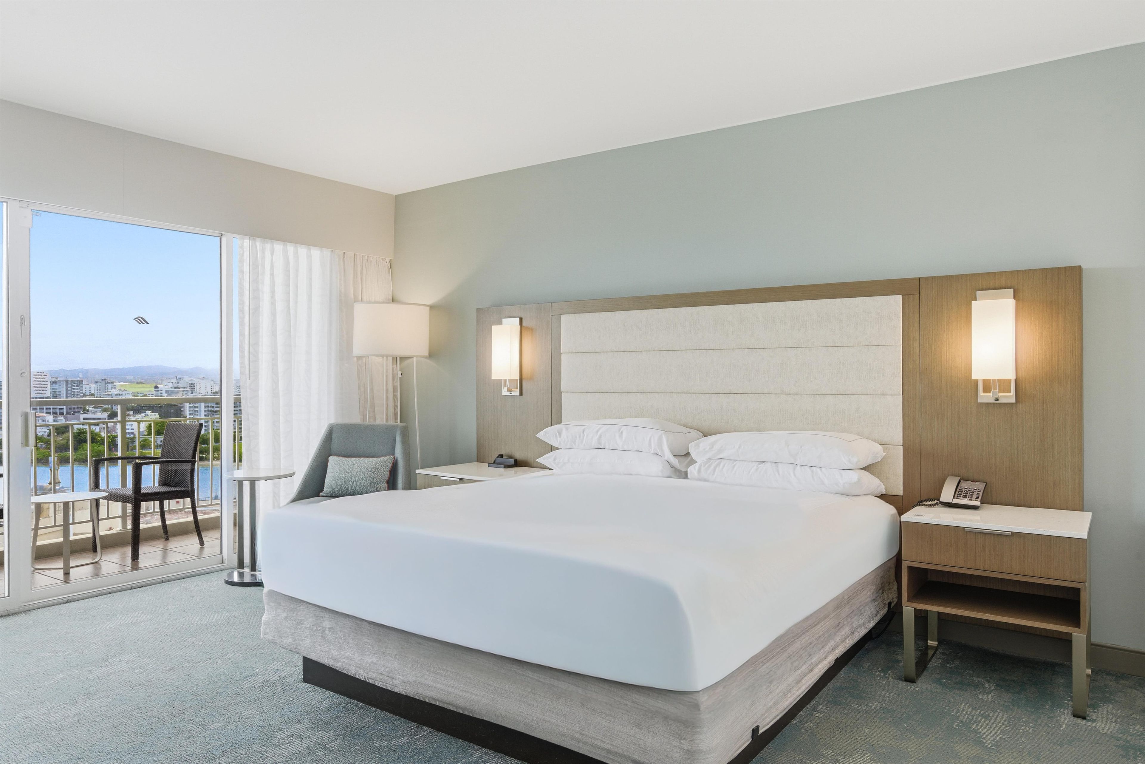 A hotel room with a large bed, a nightstand, a chair, a lamp, and a view of the city.
