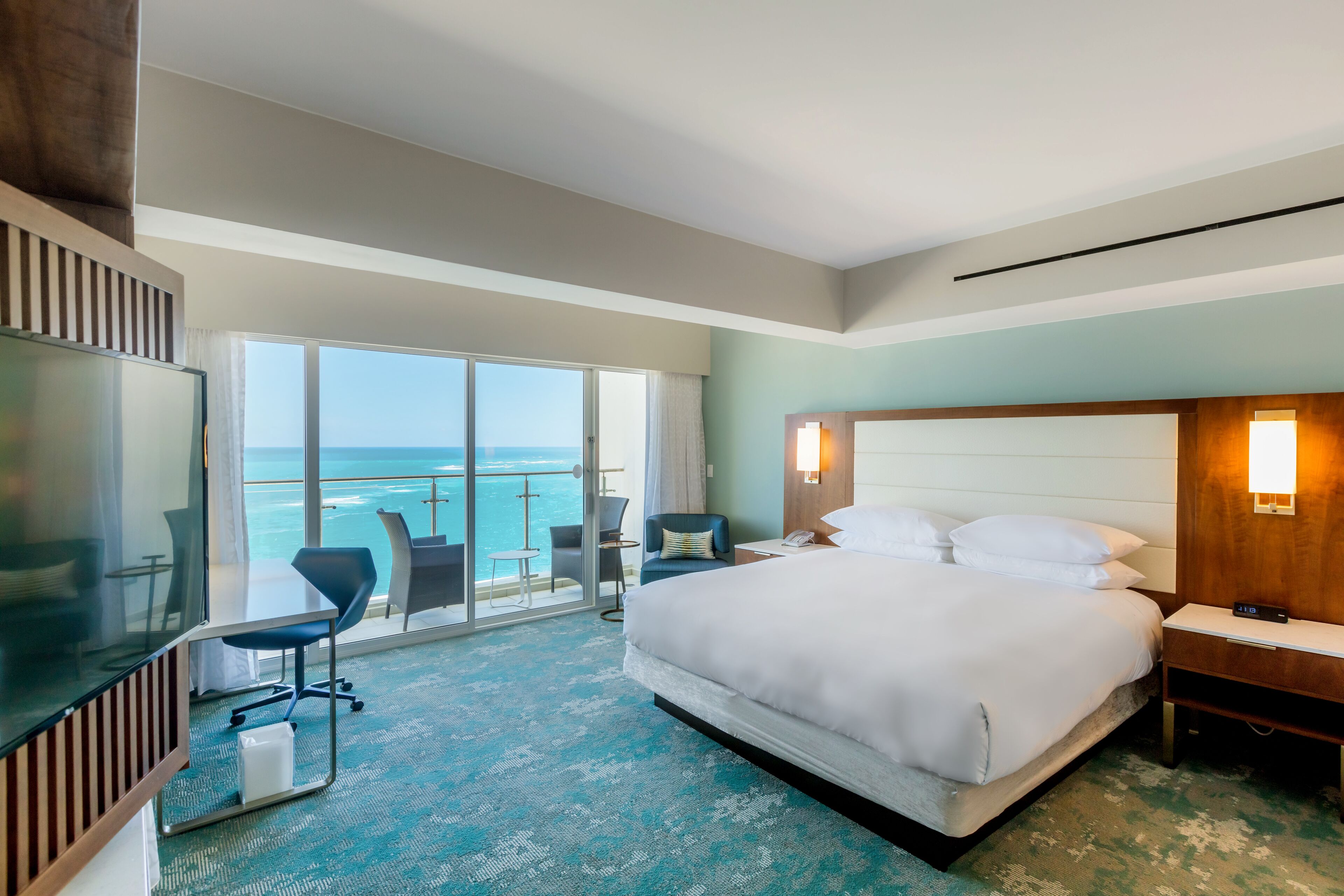 A hotel room with a large bed, a desk, a chair, and a TV. There is a balcony with a view of the ocean.