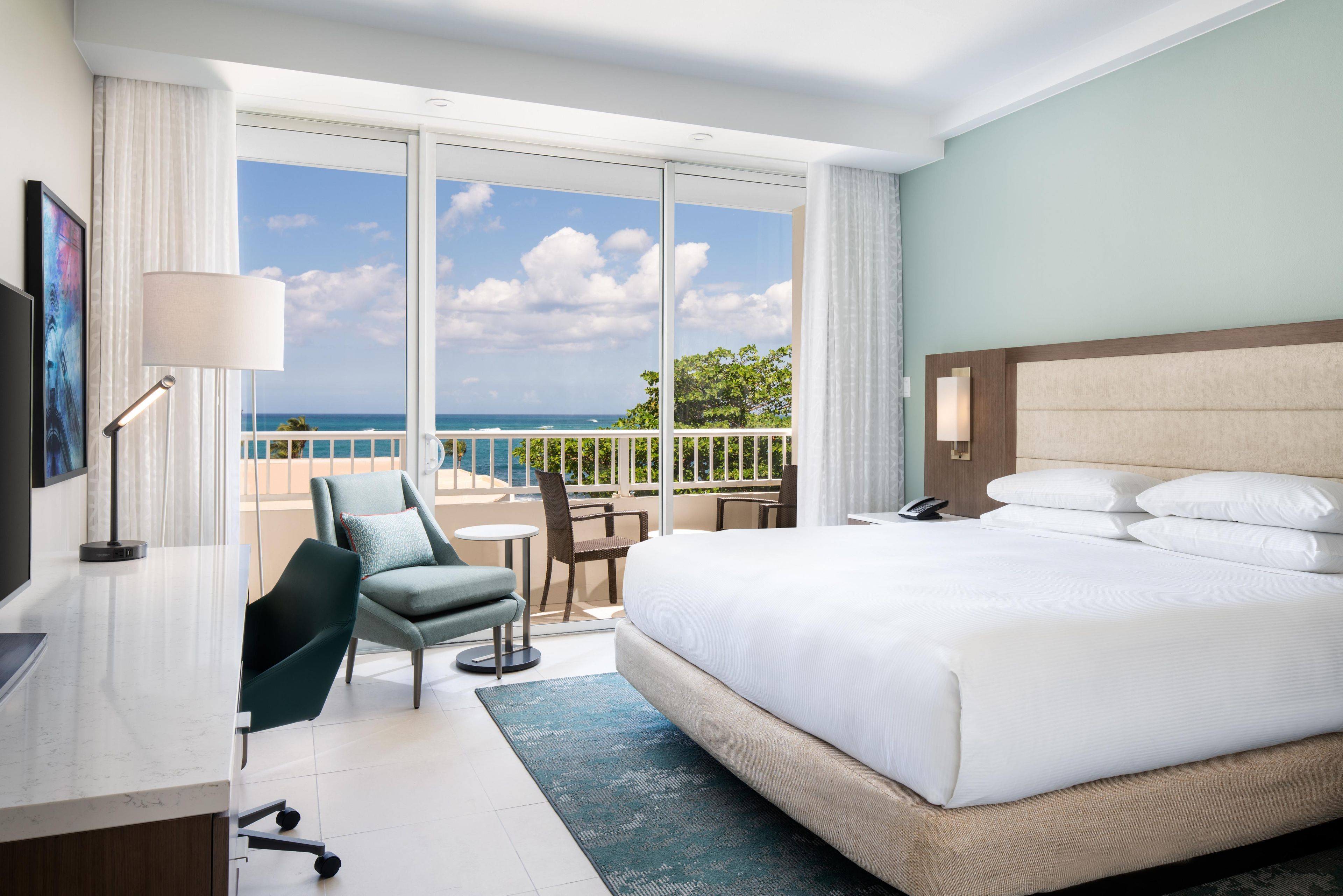 Habitación de hotel con una cama grande, un escritorio con lámpara, una silla, una mesita y vistas al océano a través de amplios ventanales.