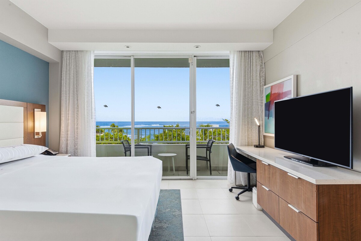 Habitación de hotel con cama, televisión, escritorio y balcón con vista al océano.