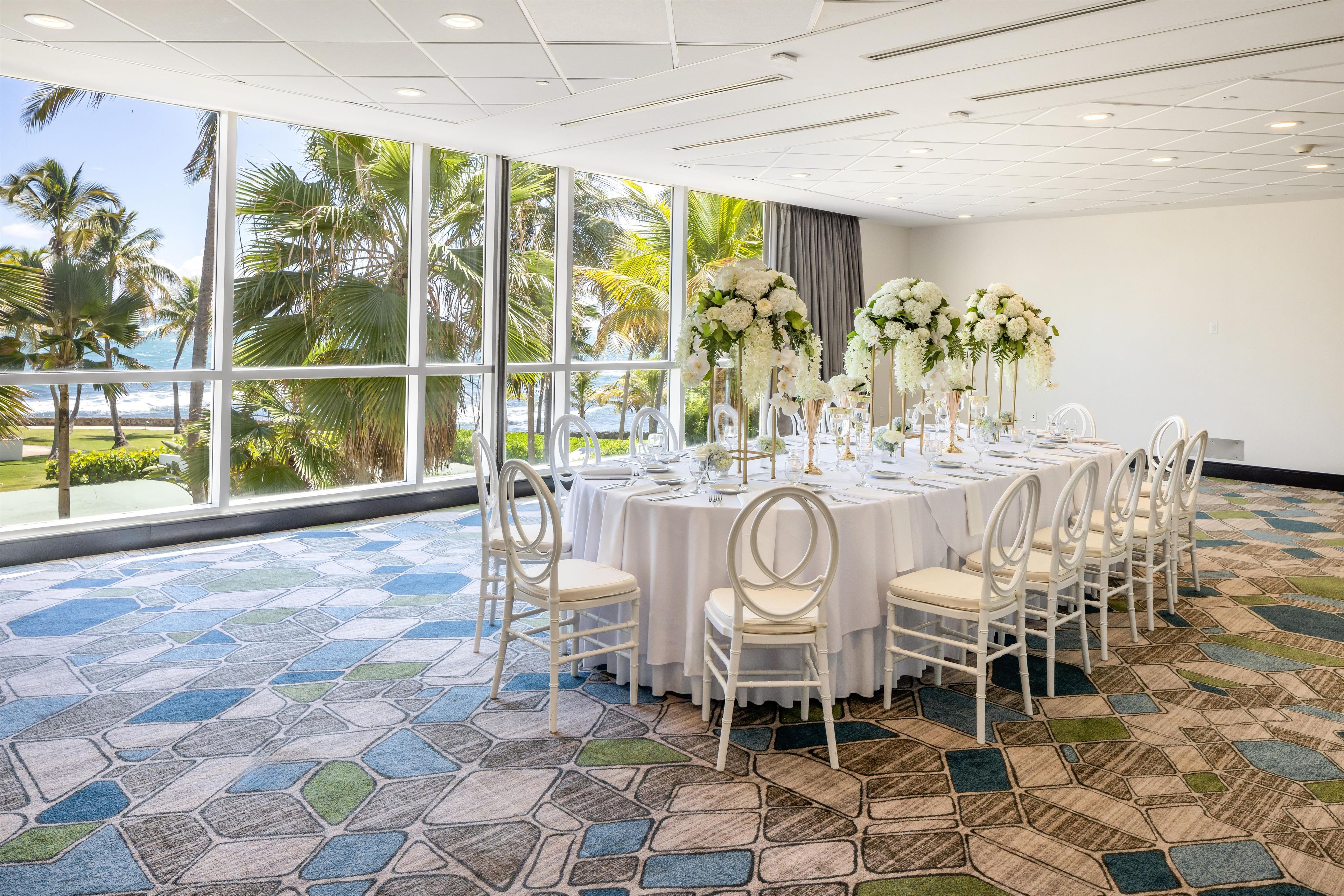 A conference room with a large window overlooking a beach, round tables set with white tablecloths and chairs, and floral centerpieces.