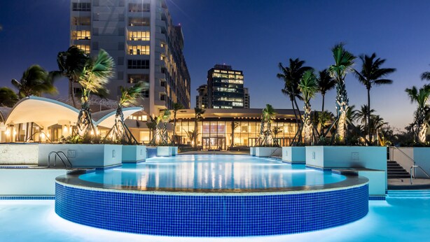 A swimming pool with a blue-tiled edge, surrounded by palm trees and modern buildings in the background. - Caribe Hilton (San Juan)