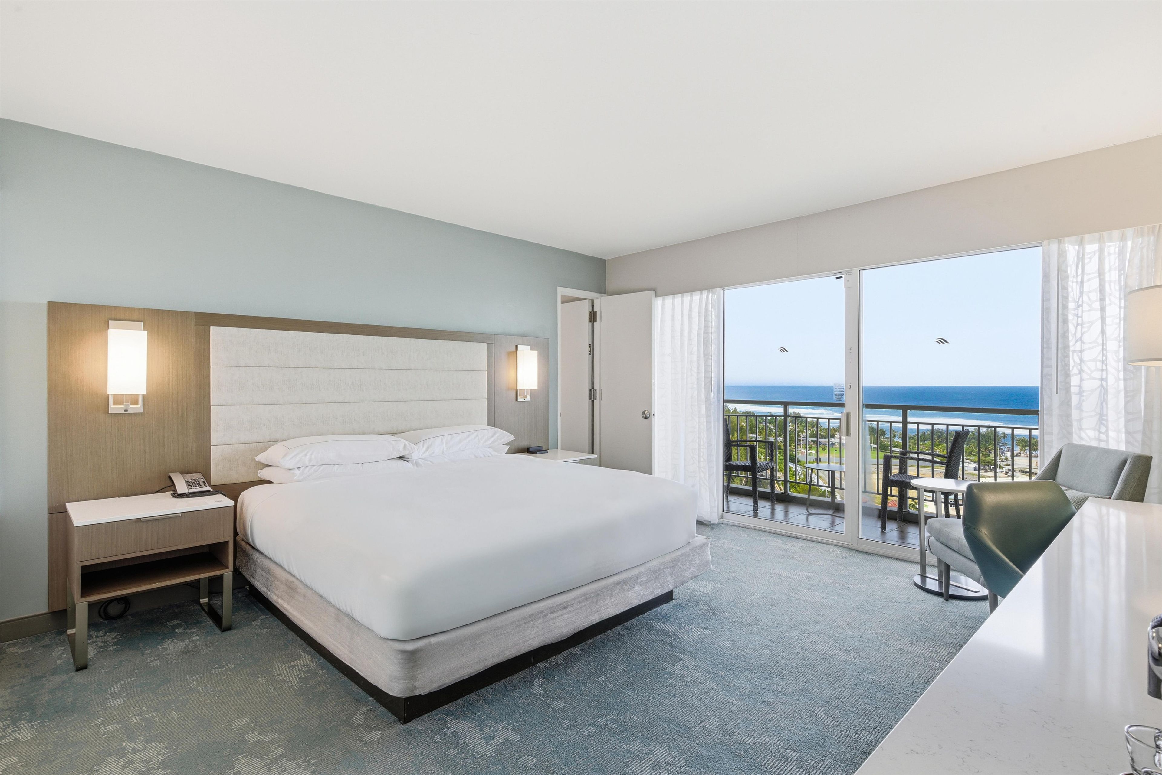 Een hotelkamer met een bed, nachtkastje, lamp en uitzicht op de oceaan vanaf een balkon.