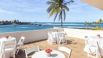 Una terraza con mesas y sillas blancas, con vistas a una playa con palmeras y el océano.