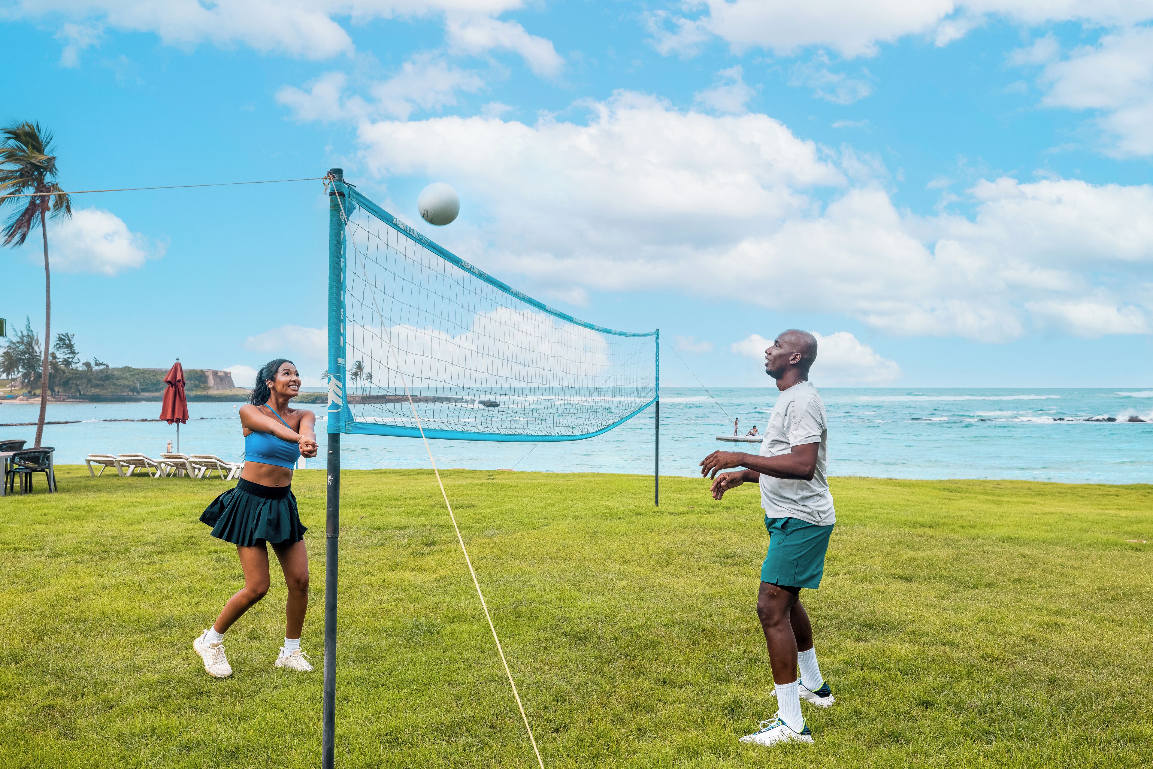 Dos personas jugando voleibol en un campo de césped con vista al océano y palmeras al fondo.