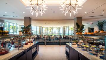 A spacious hotel dining area with a buffet setup, large windows, and a modern chandelier.