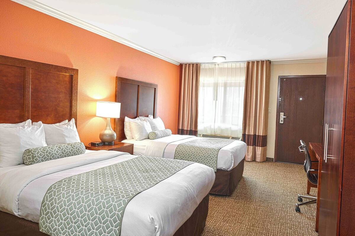 A hotel room with two beds, a nightstand with a lamp, a desk with a chair, and a wardrobe.