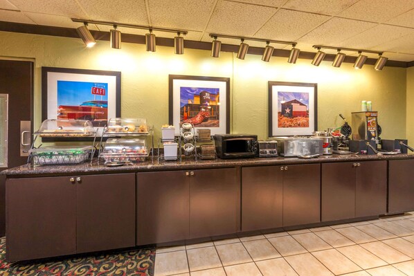 A buffet counter with various food items, a coffee machine, and a microwave. There are framed pictures on the wall and a refrigerator with beverages.