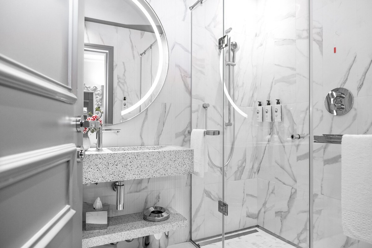 A modern bathroom with a marble shower, a round mirror, and a granite countertop.