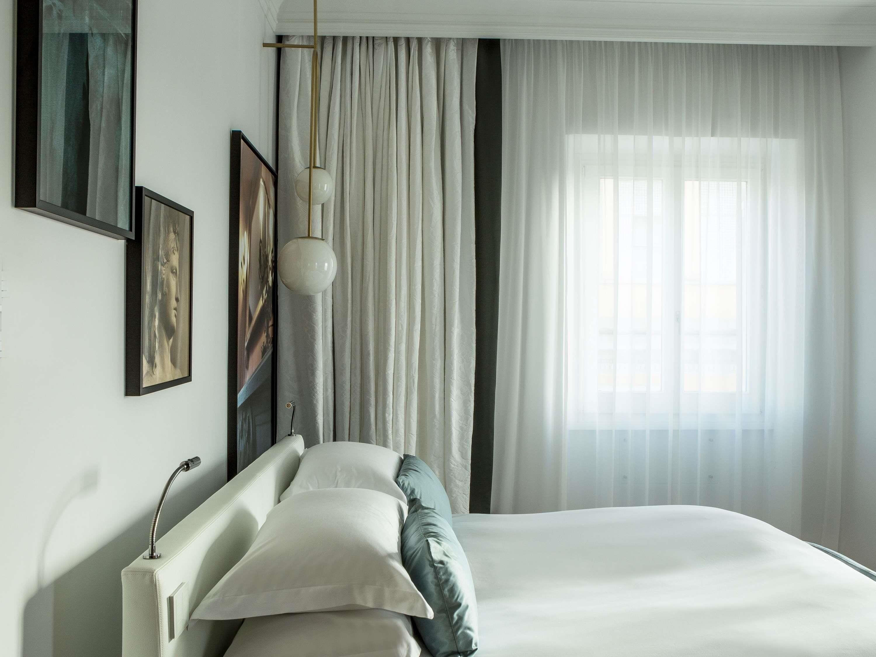 Quarto com cama, luminária pendente e janela com cortinas finas.