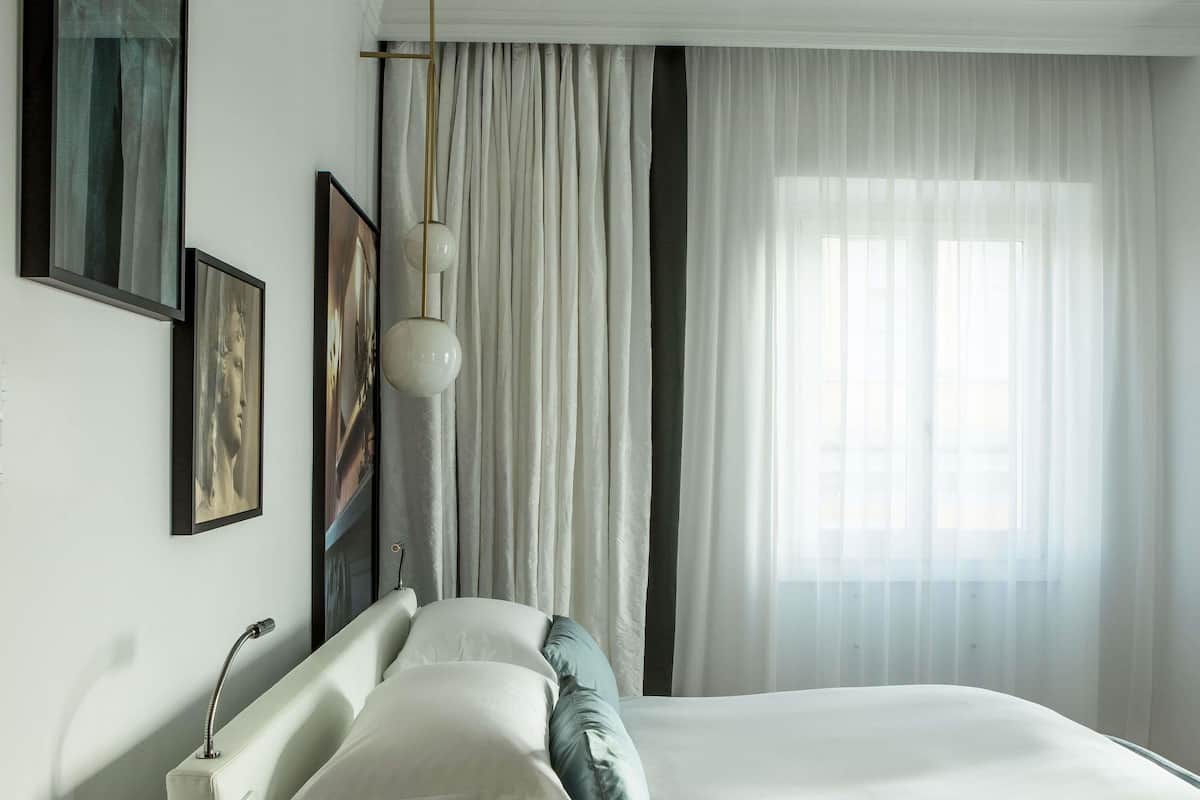 A bedroom with a bed, curtains, a hanging light fixture, and framed artwork on the wall.