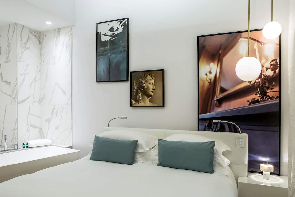 A modern bedroom with a marble wall, a bed with two pillows, a nightstand with a lamp, and artwork on the wall.