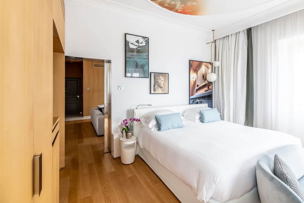 A modern hotel room with a large bed, a bedside table with a lamp, a small side table with a plant, and a wardrobe.