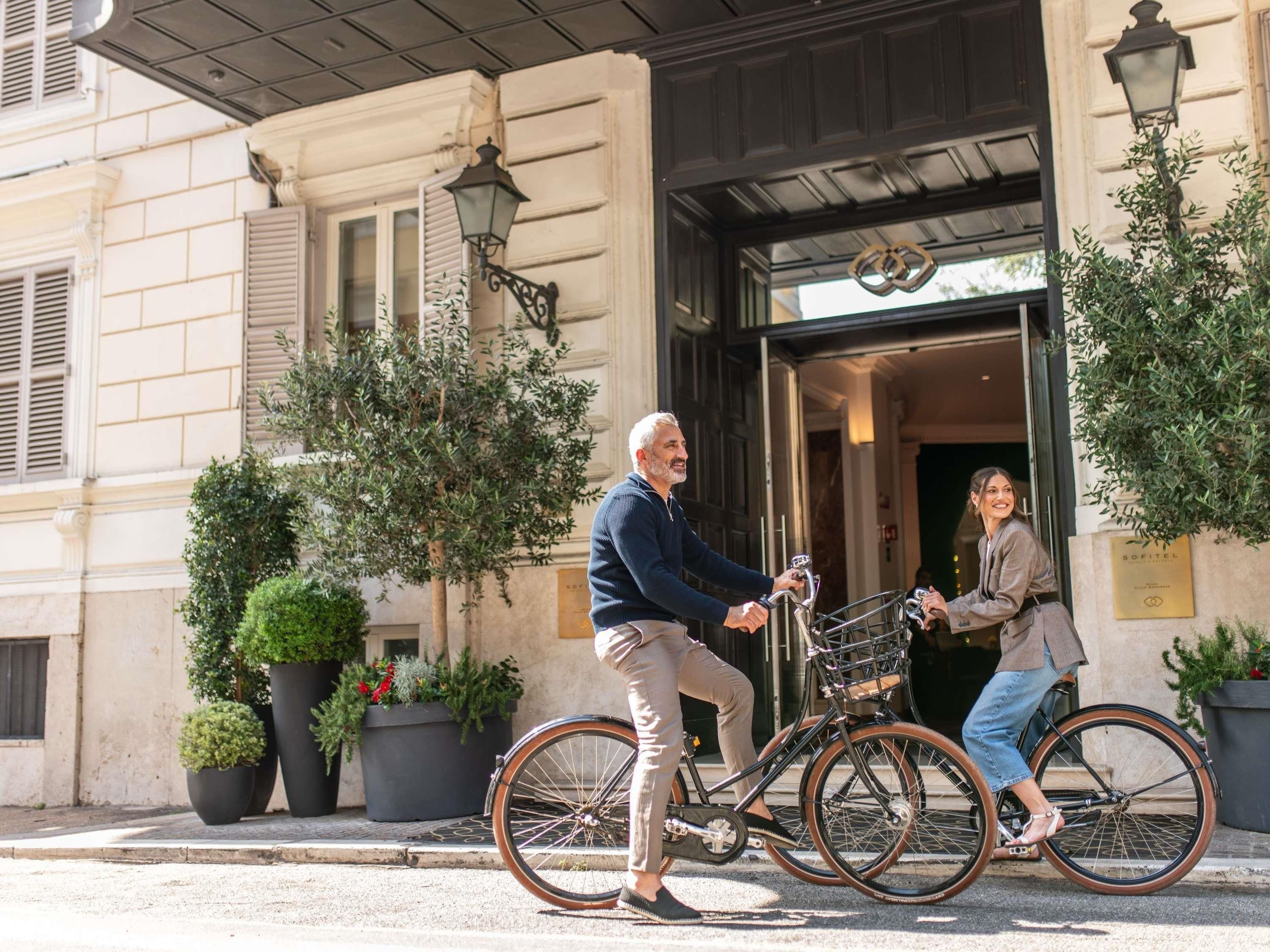 Un home amb una bicicleta i un cistella, una dona caminant al seu costat, i l'entrada d'un edifici amb plantes en testos i un rètol.