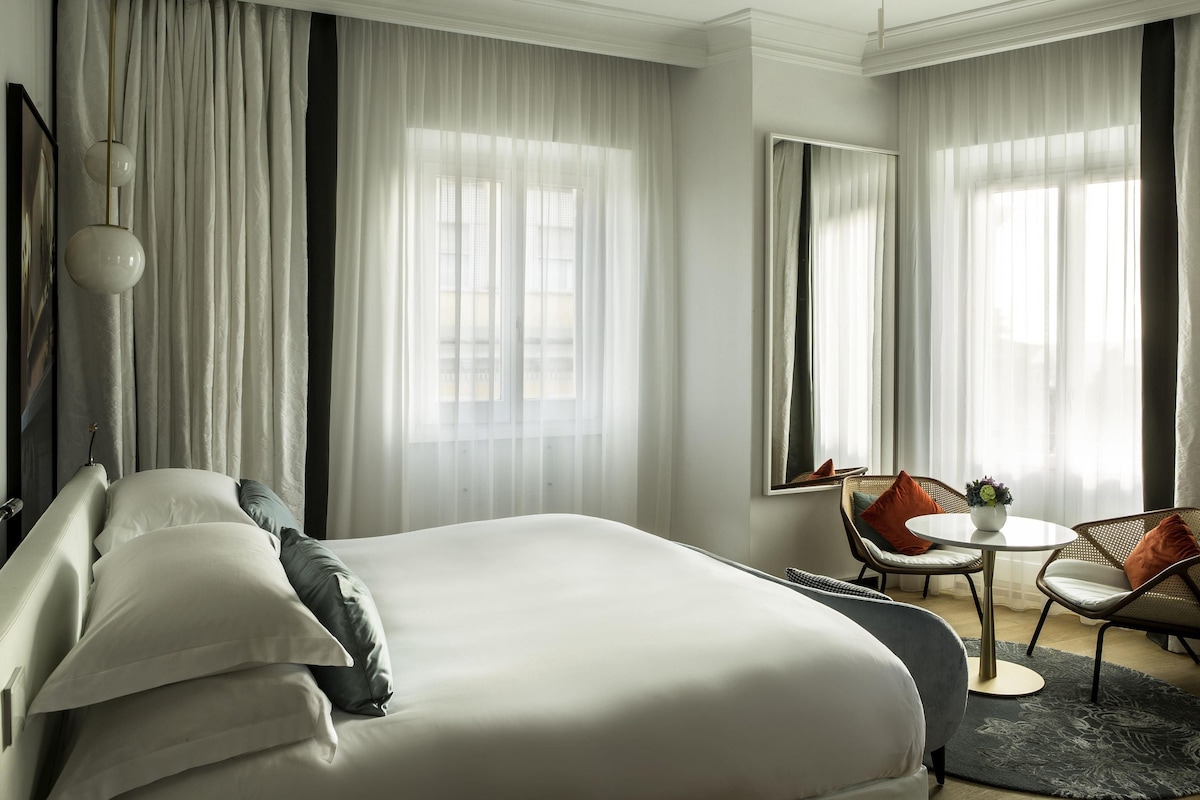 A modern hotel room with a large bed, a sitting area with a table and chairs, and large windows with sheer curtains.