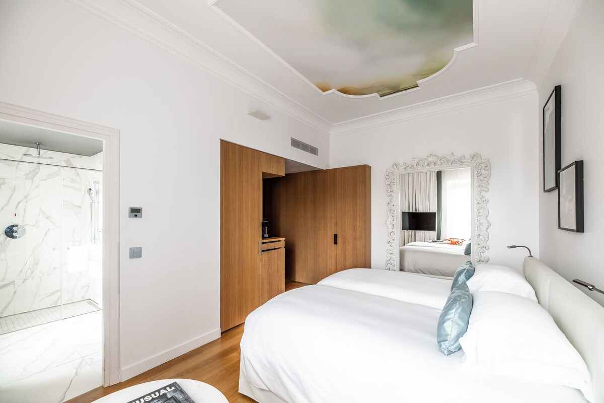 A modern hotel room with a large bed, a wooden wardrobe, a bathroom with a marble shower, and a ceiling with a unique design.