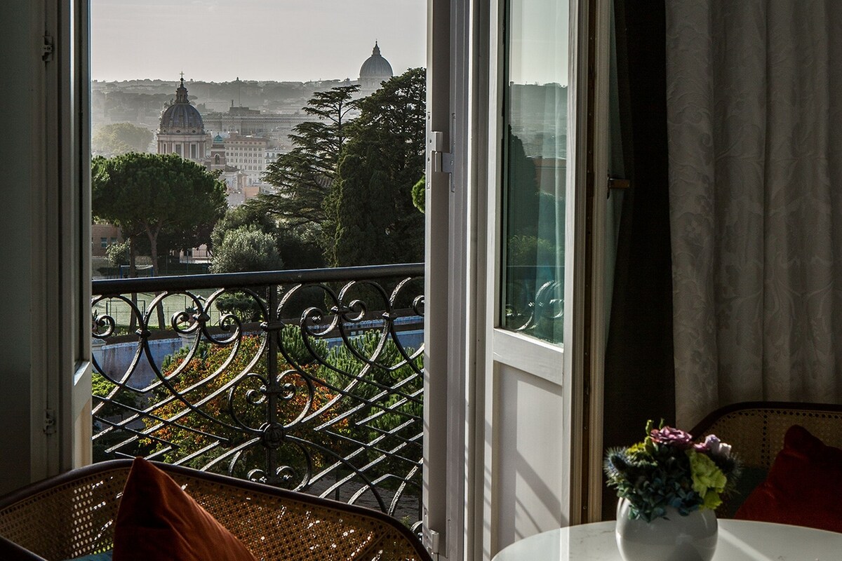 A balcony with a view of a cityscape, including a dome building, a chair with a cushion, a small table with a vase, and a curtain.