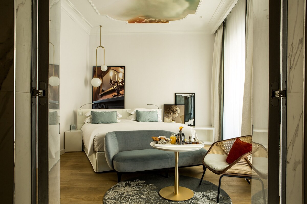 A modern hotel room with a bed, bedside table, and a small round table with a tray of breakfast items.